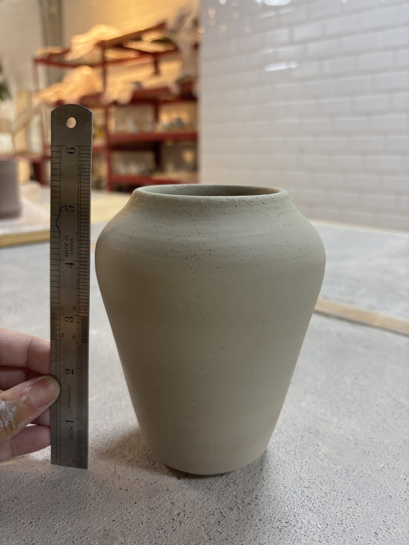 A hand is holding a ruler next to a clay vase on a table, measuring its height.