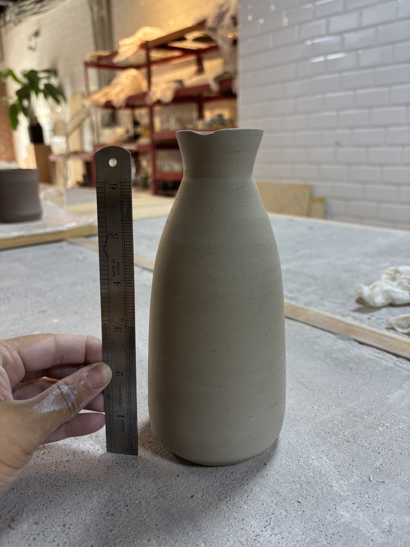 A hand holds a ruler next to an unfinished ceramic vase on a worktable in a pottery studio.