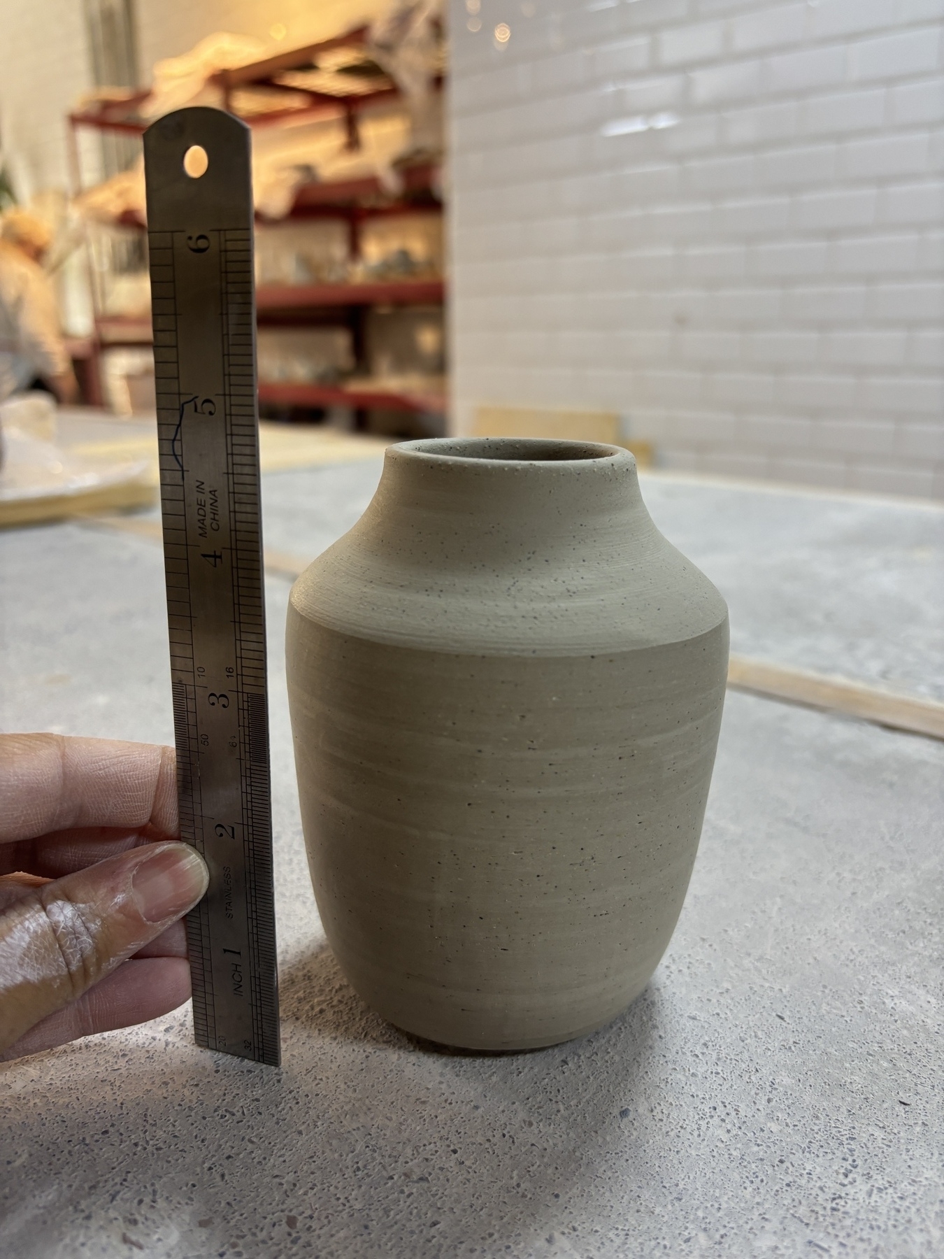 A handmade ceramic vase is being measured with a ruler inside a workshop setting.